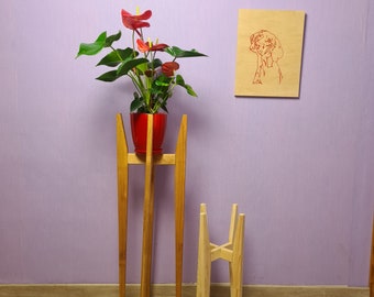 May include: A wooden plant stand with three legs and a red planter with a pink flower. The stand is in front of a light purple wall with a wooden picture frame hanging on the wall.