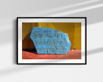 Op de afbeelding: Een blauwe steen met daarin gegraveerde Tibetaanse schrifttekens, tegen een gele muur. De steen ligt op een rood oppervlak.