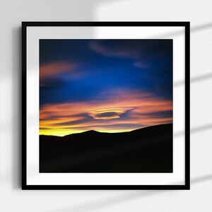 May include: A framed photograph of a sunset over a mountain range. The sky is a vibrant mix of orange, yellow, and blue, with a ring of clouds in the center. The mountains are silhouetted against the sky.