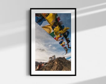 May include: Framed print depicting a mountain scene with a fortress and vibrant prayer flags. The flags, in yellow, blue, red, and green, are strung across the sky. The image is set against a backdrop of a cloudy sky.