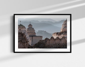 May include: Framed print depicting a mountain landscape with ancient structures. The image shows weathered stone buildings with detailed features, set against a backdrop of layered mountain ranges under a hazy sky. The print is in a black frame.