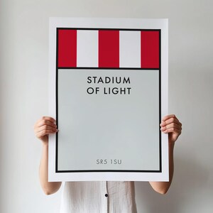 May include: A rectangular wall art print featuring a top section with alternating red and white vertical stripes. Below this, a light gray field displays the black text STADIUM OF LIGHT in the center and the alphanumeric code SR5 1SU at the bottom.