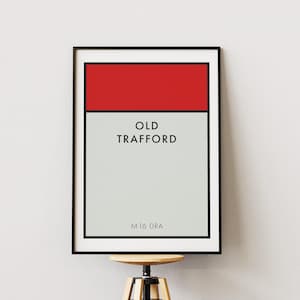 May include: A framed rectangular wall art print featuring a red horizontal bar at the top and a light gray background below. The text OLD TRAFFORD is printed in black in the center, with the alphanumeric code M16 0RA at the bottom.