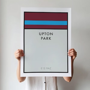 May include: A rectangular wall art print featuring a maroon and light blue horizontal stripe at the top, a light gray background, and black text that reads UPTON PARK and E13 9AZ, all enclosed within a thick black border on a white paper sheet.