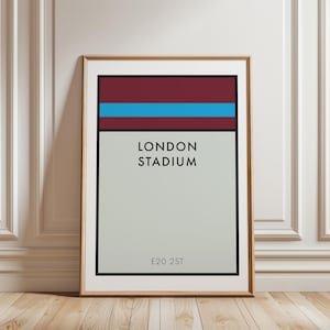 May include: A framed rectangular print featuring a minimalist graphic design with a maroon top section, a horizontal light blue stripe, and a large light gray area. The text LONDON STADIUM is centered in black, with E20 2ST printed at the bottom.
