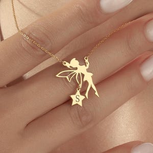 May include: A gold necklace with a fairy pendant and a star charm. The fairy has outstretched arms and wings, and the star charm has the letter "S" engraved on it. The necklace is suspended from a fine chain.