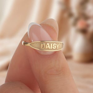 Könnte beinhalten: Ein goldfarbener Ring mit dem eingravierten Namen "DAISY" auf einer strukturierten rechteckigen Platte. Der Ring wird zwischen Fingern gehalten, was das personalisierte Schmuckstück zeigt.
