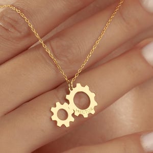 May include: A gold-colored necklace featuring two gear-shaped pendants. The larger gear has the name "Hector" engraved on it. The necklace is displayed against a skin-toned background, highlighting the jewelry's design.