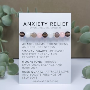Könnte beinhalten: Eine weiße Karte mit dem Text "Anxiety Relief Crystal Healing Jewellery" und einer Beschreibung der heilenden Eigenschaften von Achat, Rauchquarz, Mondstein und Rosenquarz. Die Karte enthält auch ein Armband aus diesen Kristallen.