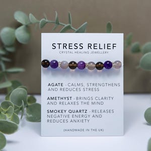 Peut inclure: Un bracelet en perles de couleur violet, marron et gris avec des perles argentées. Le bracelet est étiqueté "Stress Relief Crystal Healing Jewellery" et liste les pierres utilisées : Agate, Améthyste et Quartz fumé. Le bracelet est fait à la main au Royaume-Uni.
