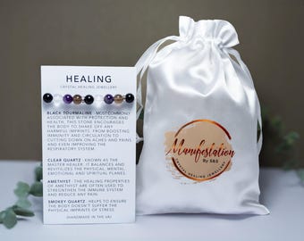 May include: A white satin pouch with the text "Manifestation by SBS Crystal Healing Jewellery" printed on it. A white card with the text "Healing Crystal Healing Jewellery" and descriptions of various crystals, including Black Tourmaline, Clear Quartz, Amethyst, and Smokey Quartz.