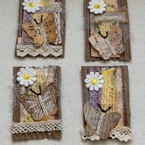 Könnte beinhalten: Vier handgefertigte rechteckige Kunstwerke. Jedes zeigt einen Schmetterling aus Notenblättern, eine weiße Gänseblümchenblüte und Spitzenbesatz. Die Hintergründe haben eine Mischung aus Texturen und Farben, darunter Lila, Gelb und Braun.
