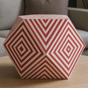 Peut inclure: Objet décoratif géométrique à motifs rouges et blancs. L'objet a une forme à facettes multiples avec un motif de losanges répétitif. L'objet est posé sur une surface en bois brun clair.