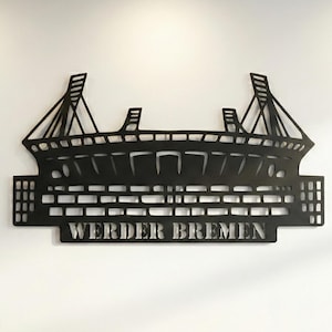Puede incluir: Arte mural de metal negro que representa el contorno de un estadio, con el texto "WERDER BREMEN" en la parte inferior. El diseño del estadio presenta detalles arquitectónicos. Montado en una pared blanca.