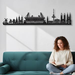 May include: Black metal wall art showing a city skyline, featuring a windmill, buildings, and a stadium. The artwork is mounted on a white wall above a teal sofa. A person is sitting on the sofa reading a book.