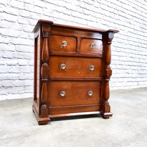 May include: A dark brown wooden chest of drawers with four drawers and crystal knobs. The chest of drawers has turned legs and a simple design.