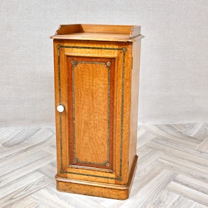 May include: A tall, rectangular wooden cabinet featuring a light brown finish with a darker wood-toned central door panel. The door is accented by a decorative border with intricate patterns and a single, small, round white knob on the left side.
