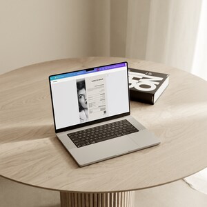 May include: A silver laptop displaying a document with the text "hello I'm Sarah" and a black and white image of a woman. A black book with white text is on a light wood table.