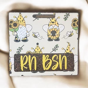 May include: A white and yellow badge with a brown border and a hole for a lanyard. The badge has a glitter background and the text "RN BSN" in yellow letters. The background features a pattern of gnomes with sunflowers and bees.