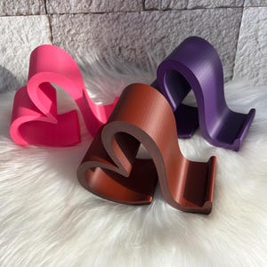 May include: Three abstract, curved sculptures in shades of pink, brown, and purple. The forms have a heart-like shape and stand on a white, fluffy surface. The sculptures appear to be made of a solid material.