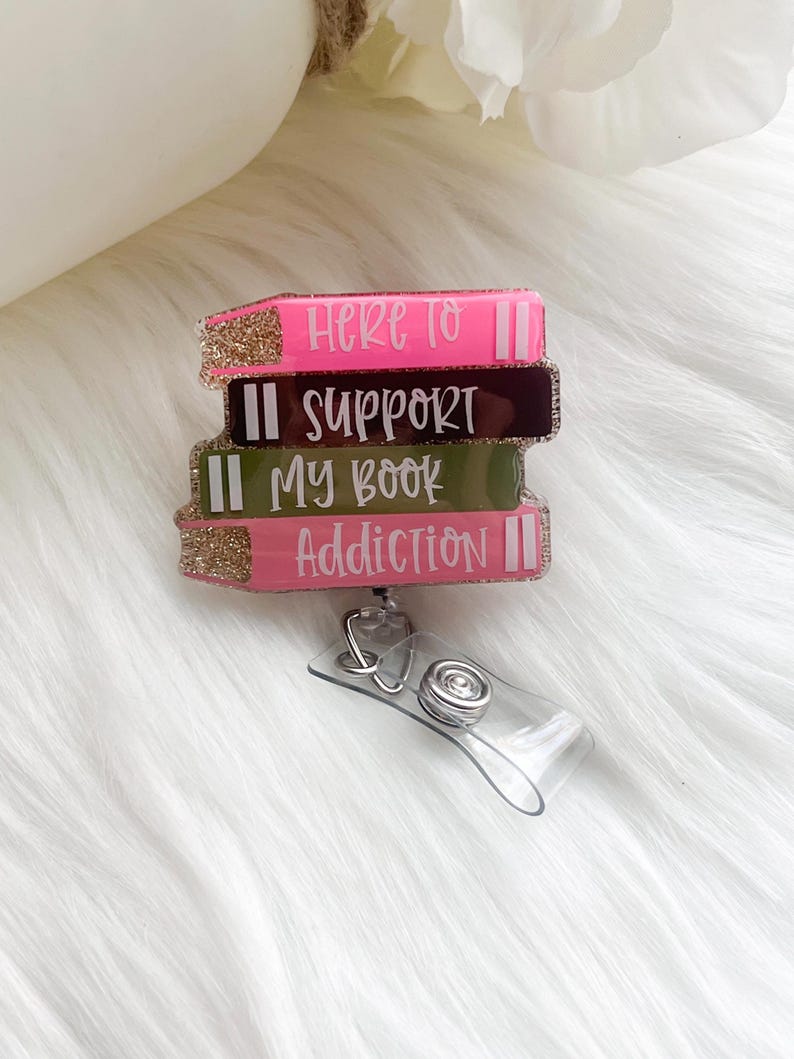 May include: A badge reel featuring a stack of four book-shaped designs. Each book is a different color: pink, dark brown, olive green, and pink. The text on the books reads "Here to Support My Book Addiction". The edges are glittery.