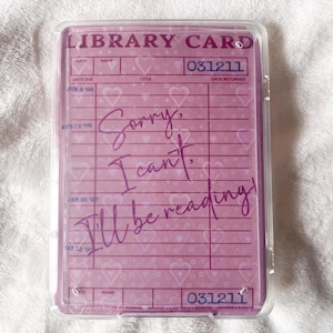 May include: A pink library card design with the text "Sorry, I can't, I'll be reading!" in a clear plastic case. The card has a heart pattern and the words "LIBRARY CARD" at the top.