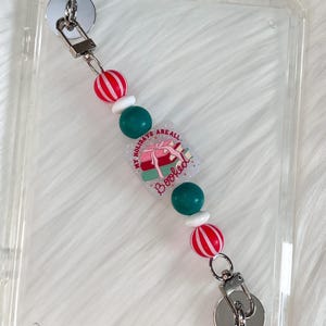 May include: A clear phone case with a beaded lanyard. The lanyard features red and white striped beads, white beads, and teal beads. A rectangular charm with the text "My Holidays Are All Booked" is also attached.