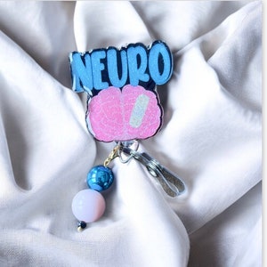 Puede incluir: Un portagafete de brillantina azul y rosa con la palabra "NEURO" y un dise&ntilde;o de cerebro. El portagafete tiene un dije de cuentas azul y blanco.