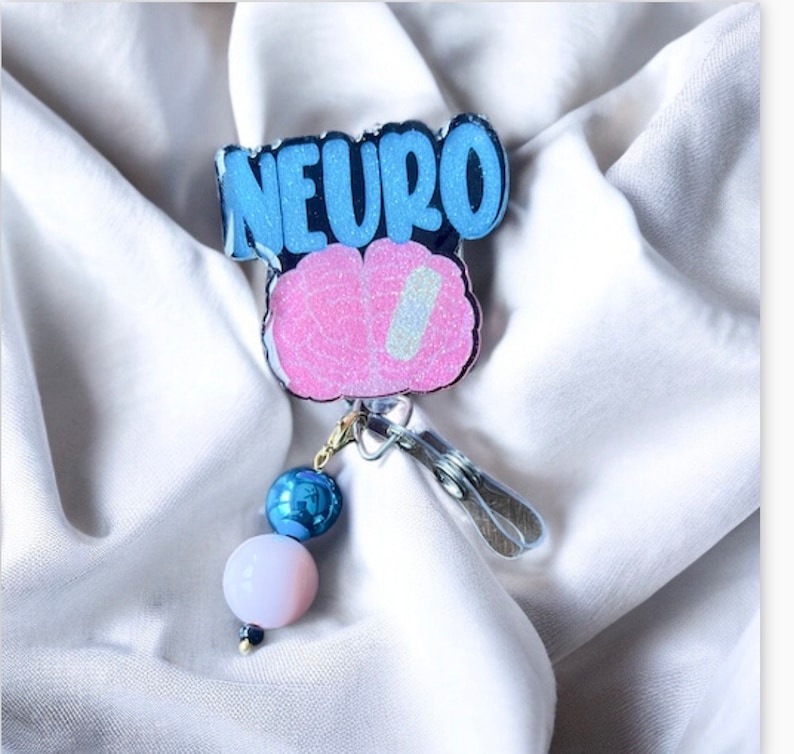 Puede incluir: Un portagafete de brillantina azul y rosa con la palabra "NEURO" y un dise&ntilde;o de cerebro. El portagafete tiene un dije de cuentas azul y blanco.