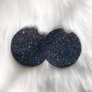 May include: Two black round coasters with iridescent glitter. The coasters are made of a durable material and are perfect for protecting surfaces from spills and scratches.