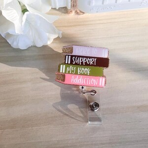 May include: A badge reel featuring a stack of colorful book-shaped charms. Each charm has glittered edges and text that reads "Here to Support My Book Addiction". The reel has a clear plastic clip and a metal retractable cord.