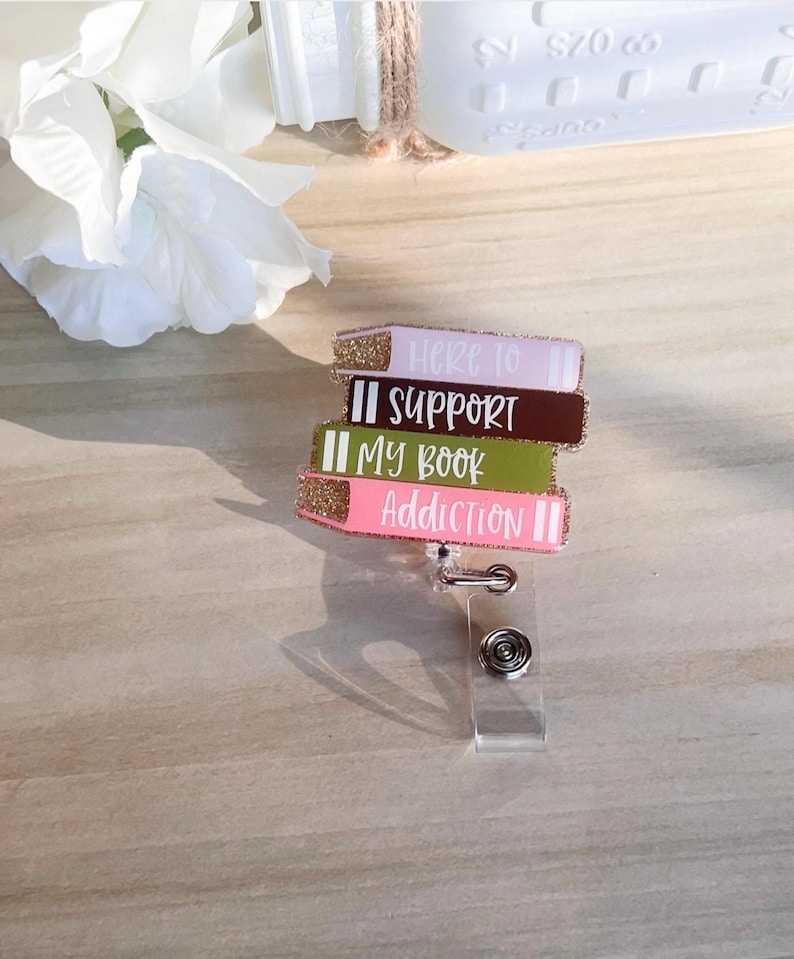 May include: A badge reel featuring a stack of colorful book-shaped charms. Each charm has glittered edges and text that reads "Here to Support My Book Addiction". The reel has a clear plastic clip and a metal retractable cord.