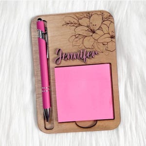 May include: A rectangular wooden note holder featuring a laser-engraved floral design at the top and the name Jennifer in script lettering. A bright pink square sticky note is attached to the center, and a hot pink pen is held in a side slot.