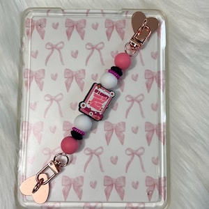 Peut inclure: Un porte-téléphone à breloques avec un fermoir et des accents en forme de cœur en or rose. La breloque comporte des perles roses, blanches et noires, et une breloque rectangulaire avec le texte "I don't take naps, I take power naps". L'étui du téléphone a un motif de nœuds roses.