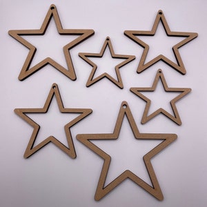 May include: Five wooden star-shaped ornaments of varying sizes. The stars are all cut out, creating a frame-like effect. The stars are a light brown color.