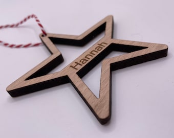 Puede incluir: Un adorno de estrella de madera con una cuerda roja y blanca para colgar. La estrella tiene un diseño recortado y el nombre "Hannah" está grabado en ella.