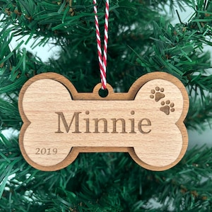 Puede incluir: Adorno navideño de madera con forma de hueso de perro, con el nombre "Minnie" y el año "2019" grabados. El adorno presenta huellas de patas y está suspendido por una cuerda a rayas rojas y blancas.