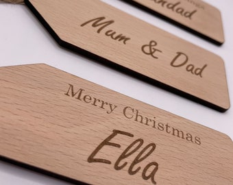 May include: Three wooden gift tags with twine attached. The tags are rectangular with rounded corners and have the following text: "Merry Christmas Ella", "Mum & Dad", and "Seasons Greetings Grandad".
