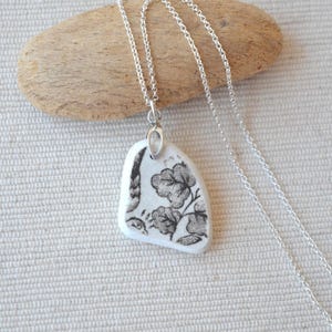May include: A silver chain necklace with a pendant made from a piece of white ceramic. The ceramic pendant has a black floral design. The necklace is displayed on a light-colored, textured surface.