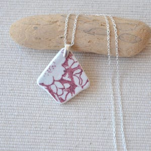 Pode incluir: Um colar com pingente em forma de losango com fundo branco e um padrão floral bordô. O pingente é preso a uma delicada corrente de prata. O colar é exibido em um pedaço de madeira flutuante.