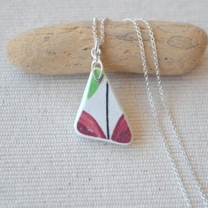 Puede incluir: Collar con colgante hecho a mano. El colgante triangular de cerámica tiene una base blanca con detalles en rojo, verde y negro. La cadena del collar es plateada. Una pieza de joyería única.