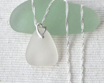 Pode incluir: Um colar com um pendente de vidro do mar branco fosco e liso, com uma forma triangular arredondada. O pendente está preso a uma delicada corrente prateada torcida por um pequeno elo metálico em forma de coração aberto no topo.