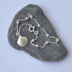 Könnte beinhalten: Ein silbernes Fußkettchen mit einem Muschel-Anhänger. Das Fußkettchen hat eine filigrane Kette mit kleinen, runden Perlen und einem Karabinerverschluss. Der Muschel-Anhänger ist aufwendig detailliert. Das Fußkettchen liegt auf einem grauen Stein.