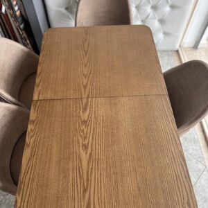 Puede incluir: Una mesa de madera con un patrón de veta de madera. La mesa es marrón y tiene un borde redondeado. La mesa está rodeada de sillas.
