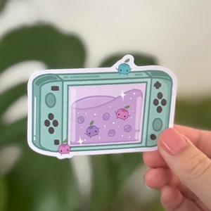 Puede incluir: Una pegatina verde y blanca de una consola de videojuegos con un slime morado y blanco en su interior. El slime tiene pequeños puntos blancos y tres pequeños personajes de dibujos animados en su interior. Los personajes son rosa, azul y morado.