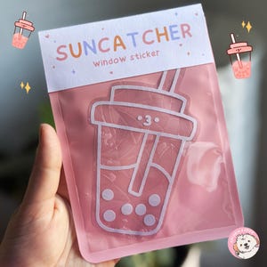 Può includere: Un adesivo per finestre rosa e bianco a forma di bicchiere di bubble tea con cannuccia e perle di tapioca. L'adesivo dice "SUNCATCHER window sticker" e ha un viso di cartone animato carino con un occhio che strizza.
