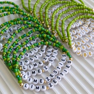 May include: A collection of beaded bracelets featuring vibrant green glass beads and white circular letter beads. Some bracelets display the text LIME ZESTIES in black, while others feature the word BESTIES in gold lettering, accented with small gold beads.