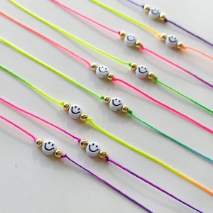 Puede incluir: Una colección de pulseras de hilo coloridas. Cada pulsera presenta una cuenta blanca con una carita sonriente y cuentas doradas. Los hilos son de varios colores brillantes, incluyendo rosa, amarillo, verde y morado. Las pulseras están dispuestas en diagonal.