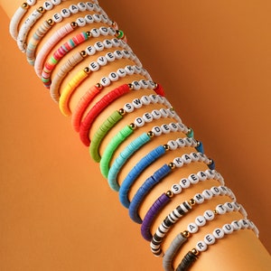 以下が含まれることがあります： 白い文字ビーズで歌のタイトルを綴った、カラフルなビーズブレスレットの積み重ね。ブレスレットは虹色のパターンで配置されており、色は白から黒まで変化しています。