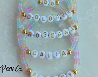 May include: Five colorful beaded bracelets with gold beads and white letter beads spelling out the names "Audrey", "Dalia", "Roseann", "Dolores", and "Bo".